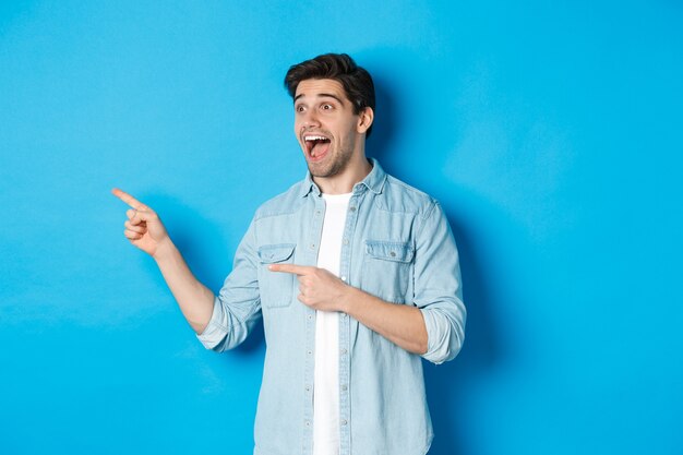 Retrato de homem bonito, olhando e apontando os dedos deixados com entusiasmo, de pé contra um fundo azul.