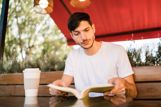 Retrato de homem caucasiano, aproveitando o tempo livre e lendo um livro enquanto está sentado ao ar livre na cafeteria. Conceito de estilo de vida.