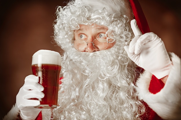 Retrato de homem com fantasia de Papai Noel - com uma luxuosa barba branca, chapéu de Papai Noel e uma fantasia vermelha em fundo vermelho do estúdio com cerveja