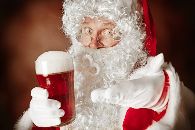 Retrato de homem com fantasia de Papai Noel - com uma luxuosa barba branca, chapéu de Papai Noel e uma fantasia vermelha em fundo vermelho do estúdio com cerveja