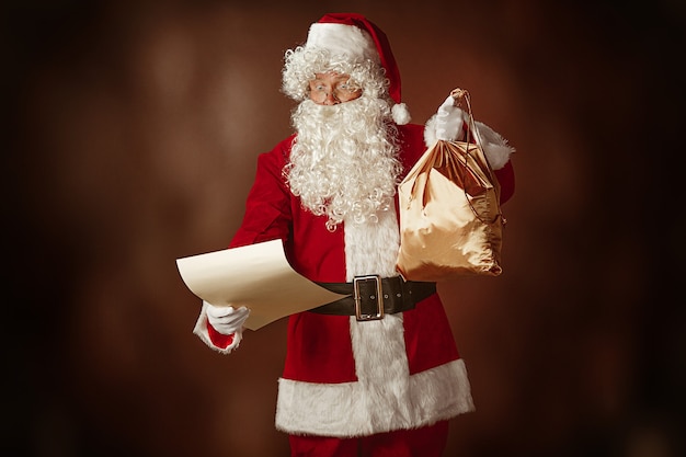 Retrato de homem com fantasia de papai noel - com uma luxuosa barba branca, chapéu de papai noel e uma fantasia vermelha lendo a carta no fundo vermelho do estúdio com presentes