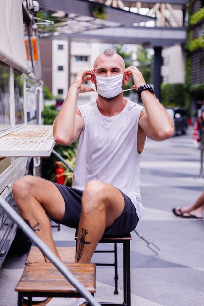 Retrato de homem com máscara médica branca na praça central da cidade se senta na cadeira no café da van