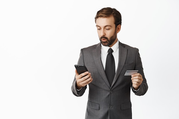 Retrato de homem corporativo olhando animado para a tela do smartphone segurando o cartão de crédito pagando pela compra de compras on-line em pé contra o fundo branco