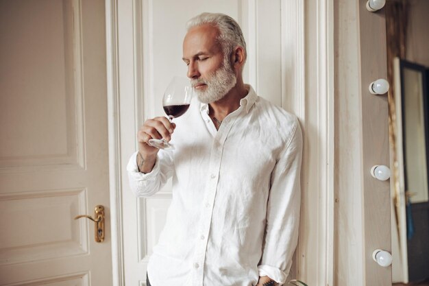 Retrato de homem de camisa branca gosta de cheiro de vinho Foto de adulto com barba grisalha em roupa leve com um copo nas mãos