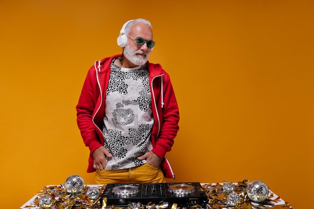 Foto grátis retrato de homem de camiseta e capuz trabalhando como dj adulto de cabelos grisalhos com barba branca em óculos de sol azuis e calças laranja posando