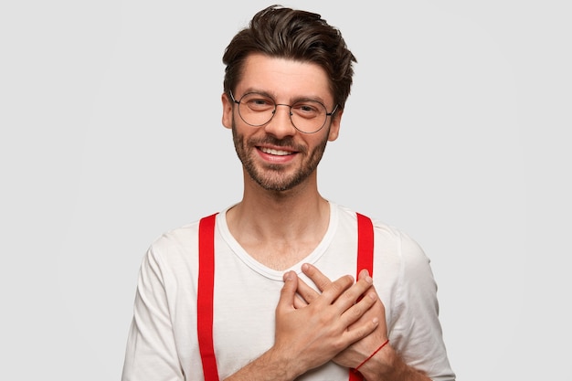 Retrato de homem feliz mantém as palmas das mãos no coração, aprecia algo com grande gratidão, vestido com traje estiloso, tem sorriso amigável, isolado sobre parede branca. Pessoas, emoções, positividade