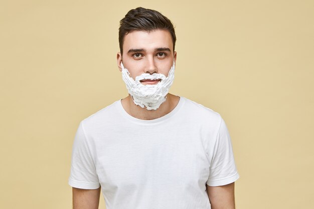 Retrato de homem jovem e bonito branco morena posar isolado com espuma de barbear branca aplicada em seu rosto, preparando a pele para fazer a barba. Rotina matinal, masculinidade, conceito de beleza e cuidado