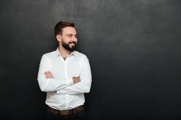 Retrato de homem positivo na camisa branca em pé com os braços cruzados e desviar o olhar sobre o espaço da cópia cinza escuro