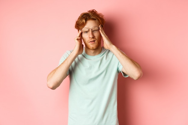 Retrato de homem ruivo de óculos tortos, tocando a cabeça e sentindo tonturas ou náuseas, tendo ressaca ou dor de cabeça, em pé sobre um fundo rosa.