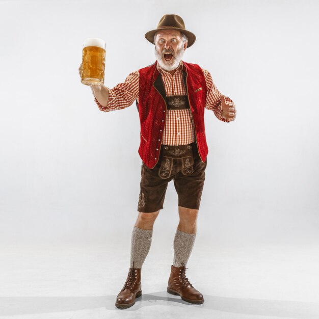 Retrato de homem sênior da Oktoberfest com chapéu, vestindo as roupas tradicionais da Baviera. Tiro completo masculino no estúdio em fundo branco. A celebração, feriados, conceito de festival. Bebendo cerveja.