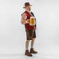 Foto grátis retrato de homem sênior da oktoberfest com chapéu, vestindo as roupas tradicionais da baviera. tiro completo masculino no estúdio em fundo branco. a celebração, feriados, conceito de festival. bebendo cerveja.