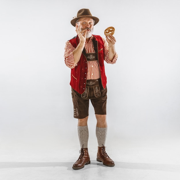 Foto grátis retrato de homem sênior da oktoberfest com chapéu, vestindo as roupas tradicionais da baviera. tiro completo masculino no estúdio em fundo branco. a celebração, feriados, conceito de festival. comer puff.