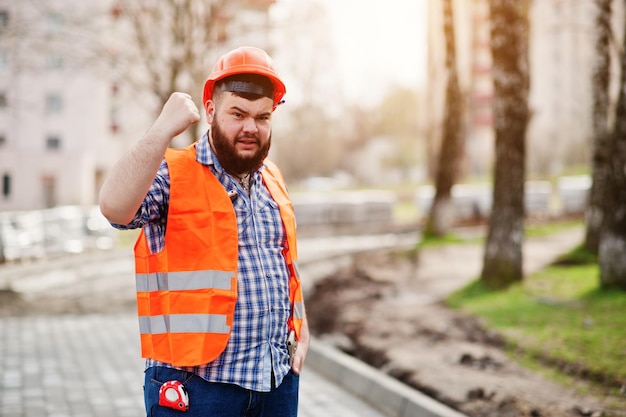 Retrato de homem trabalhador de barba com raiva terno trabalhador da construção civil em capacete laranja de segurança contra pavimento com mostrando os braços