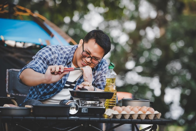 Retrato de homem viajante asiático óculos bife de porco fritando churrasco em assar frigideira ou panela em um acampamento Cozinhar ao ar livre viajando acampamento conceito de estilo de vida