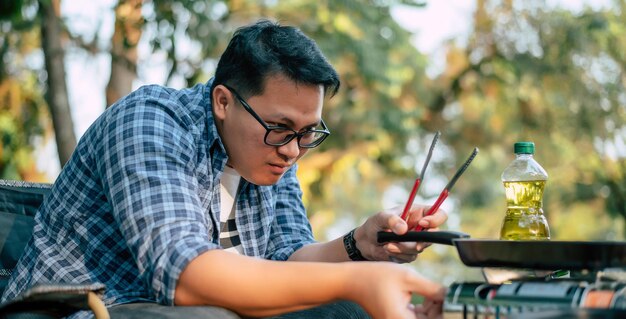 Retrato de homem viajante asiático óculos bife de porco fritando churrasco em assar frigideira ou panela em um acampamento Cozinhar ao ar livre viajando acampamento conceito de estilo de vida