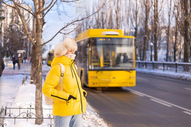 Retrato de inverno de uma mulher feliz em um casaco amarelo quente e chapéu russo siberiano esperando o ônibus em uma rua da cidade cheia de neve