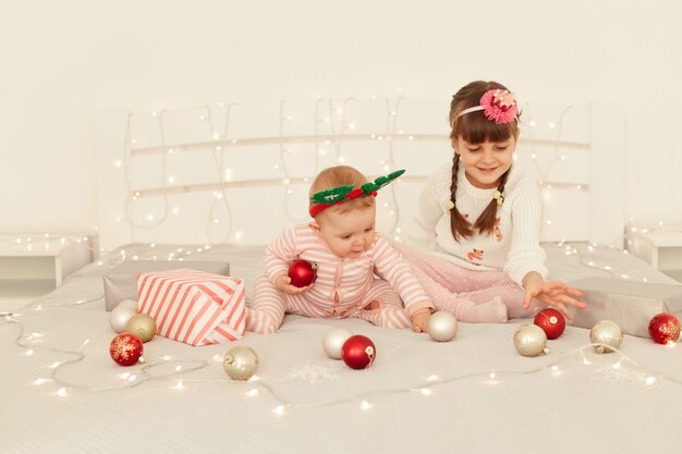 Retrato de irmãs encantadoras, lindas meninas brincando com bolas de Natal na cama decorada com luzes, crianças vestindo trajes casuais, feliz ano novo.