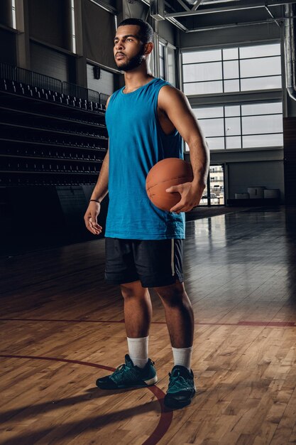 Retrato de jogador de basquete preto detém uma bola sobre um aro em um salão de basquete.