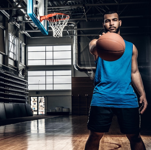 Retrato de jogador de basquete preto detém uma bola sobre um aro em um salão de basquete.