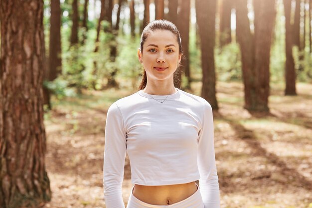 Retrato de jovem adulto feminino atraente de cabelos escuros em roupas esportivas elegantes, posando na floresta, antes ou depois do treino