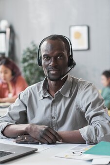 Retrato de jovem africano em fones de ouvido, olhando para a câmera enquanto está sentado à mesa no atendimento ao cliente