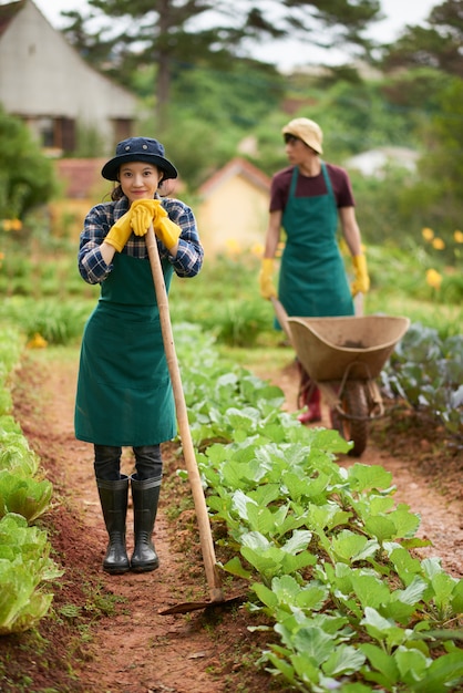 Retrato de jovem agricultor com enxada, olhando para a câmera com seu colega empurrando o vagão no fundo