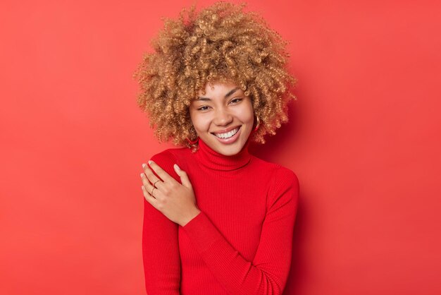 Retrato de jovem alegre mantém a mão no ombro sorri suavemente expressa expressões autênticas felizes vestidas de gola alta casual isolada sobre fundo vermelho vívido. Conceito de felicidade