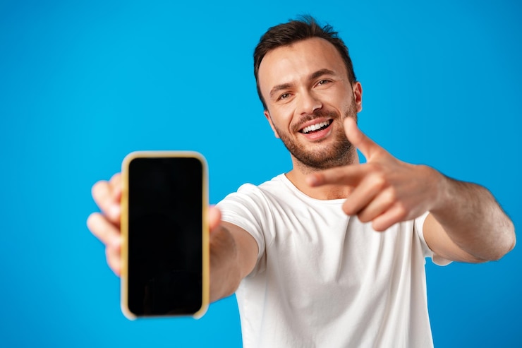 Foto grátis retrato de jovem anunciando um novo smartphone, mostrando-o para a câmera contra um fundo azul