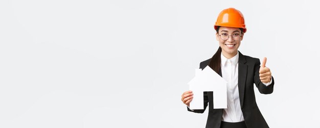 Retrato de jovem arquiteto engenheiro de construção feminino asiático em capacete de segurança e terno de negócios segurando casa maket e mostrar o polegar para cima promovendo o serviço da empresa de construção de casas