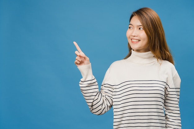 Retrato de jovem asiática sorrindo com uma expressão alegre, mostra algo incrível no espaço em branco em roupas casuais e isolado sobre a parede azul