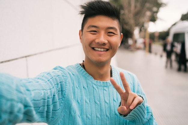 Retrato de jovem asiático parecendo confiante e tomando uma selfie em pé ao ar livre na rua.