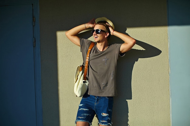 Retrato de jovem atraente sorridente moderno homem elegante em pano casual no chapéu em óculos de sol em pé perto da parede