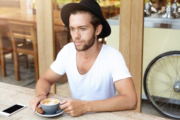 Retrato de jovem barbudo sério com chapéu da moda tomando cappuccino com biscoito enquanto relaxa no café sozinho