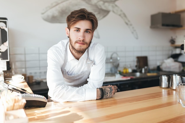 Retrato de jovem barista em pé no balcão no restaurante homem de avental e camisa branca, inclinando as mãos no balcão, olhando para a câmera no trabalho no café