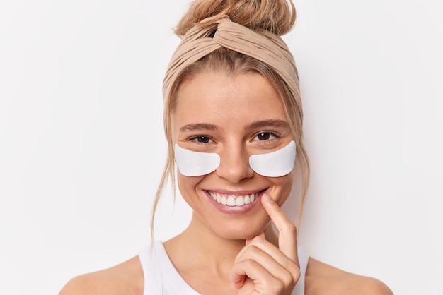 Retrato de jovem bonita e feliz aplica manchas de beleza sorrisos amplamente reduz bolsas nos olhos ou rugas mostra efeito de pele perfeita usa bandana tinha cabelo penteado isolado sobre fundo branco