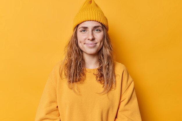 Retrato de jovem bonita olha diretamente para a câmera com um sorriso suave no rosto se sente satisfeito usa chapéu e jumper isolado sobre poses de fundo amarelo vívido para fazer memorabe shot