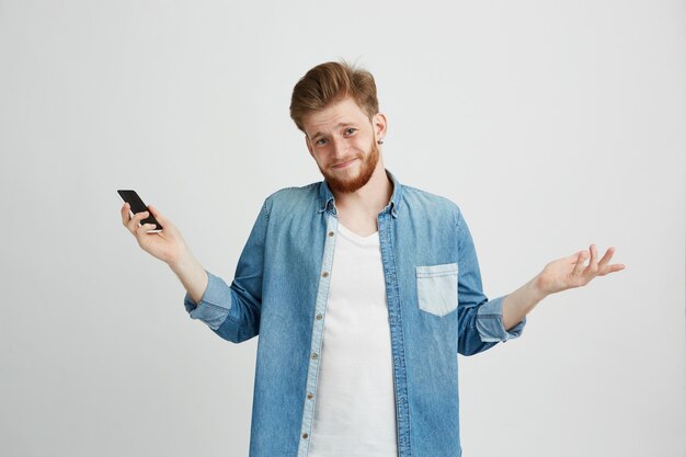 Retrato de jovem bonitão segurando o telefone inteligente, encolher os ombros, olhando para a câmera.