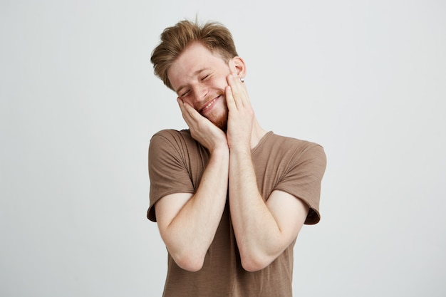 Retrato de jovem bonito bonito sorrindo com as mãos nas bochechas e olhos fechados.