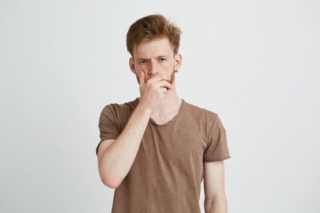 Retrato de jovem bonito brutal com barba, franzindo a testa, segurando a mão na boca.