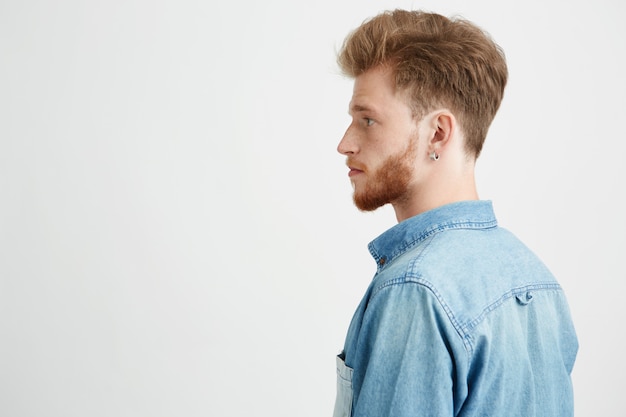 Retrato de jovem bonito com barba, vestindo a camisa de brim em pé no perfil.