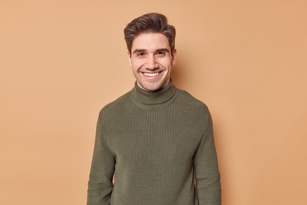 Retrato de jovem bonito com sorrisos de cabelo escuro expressa alegremente emoções positivas vestido com gola rulê casual ouve bons nes isolados sobre fundo marrom feliz em conhecer o velho melhor amigo