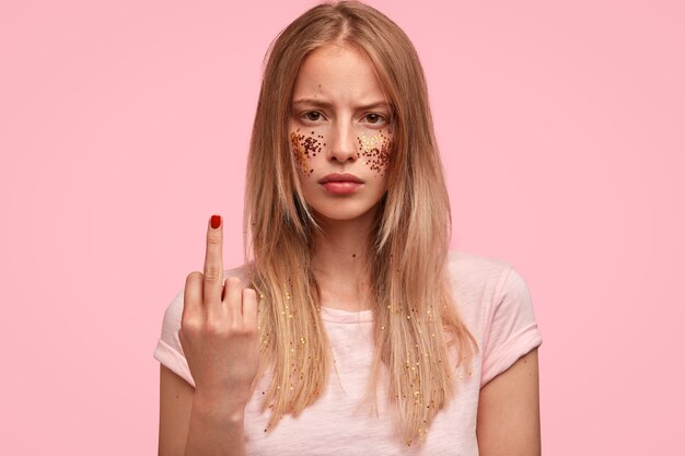 Retrato de jovem caucasiana descontente mostra dedo médio, tem brilhos nas bochechas, está desanimada, discute com alguém, usa camiseta casual rosa claro em um tom com parede