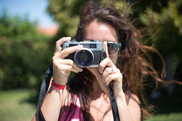 Retrato de jovem com câmera. Linda mulher de cabelos escuros em óculos de sol, segurando uma câmera fotográfica à moda antiga. Lazer, amizade, conceito de festa