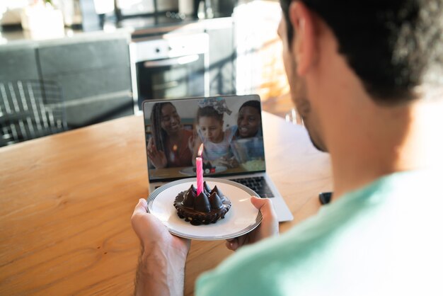 Retrato de jovem comemorando aniversário em uma chamada de vídeo em casa com o laptop e um bolo. Novo conceito de estilo de vida normal.