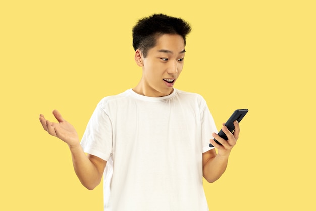 Retrato de jovem coreano. Modelo masculino em camisa branca. Usando smartphone para apostas, leitura de notícias ou conversas. Conceito de emoções humanas, expressão facial.