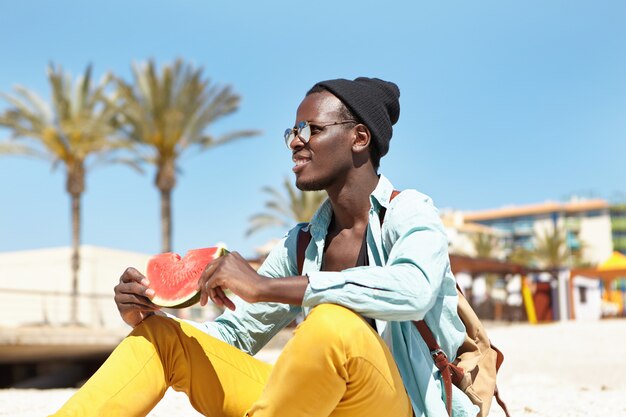 Retrato de jovem despreocupado de pele escura em elegantes chapéus e óculos de sol relaxantes na praia com uma fatia de melancia fresca e suculenta, admirando o mar azul calmo durante as férias na cidade turística