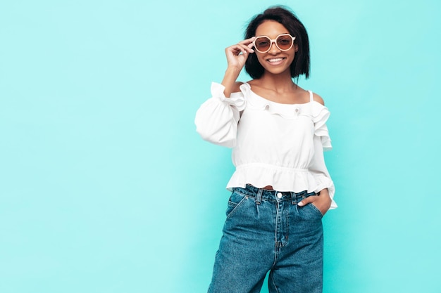 Foto grátis retrato de jovem e bela mulher negra modelo sorridente vestido com roupas de jeans de verão mulher despreocupada sexy posando perto da parede azul no estúdio bronzeado e alegre em óculos de sol