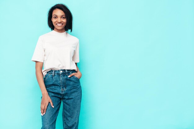 Retrato de jovem e linda mulher negra Modelo sorridente vestido com roupas de jeans de verão Mulher despreocupada sexy posando perto da parede azul no estúdio Bronzeado e alegre