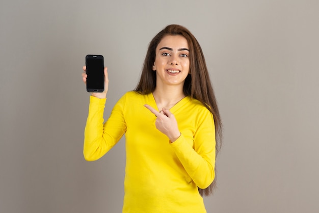 Retrato de jovem em amarelo segurando o celular na parede cinza.