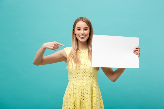 Retrato de jovem em vestido amarelo, apontando o dedo no quadro em branco do lado branco. Isolado sobre o fundo azul.